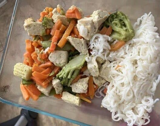 picture of a veggie stirfry with broccoli, baby corn, carrots and tofu with noodles in a glass container.