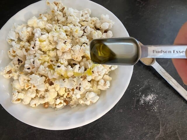 hand drizzling olive oil over plain popcorn in a bowl. There is also some salt and a measuring spoon with nutritional yeast in the background