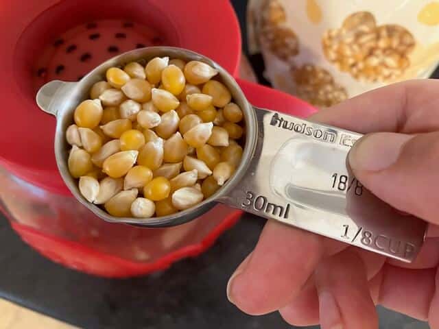 hand holding popcorn kernels in a measuring cup. There is a popcorn bag and microwave popcorn popper in the background