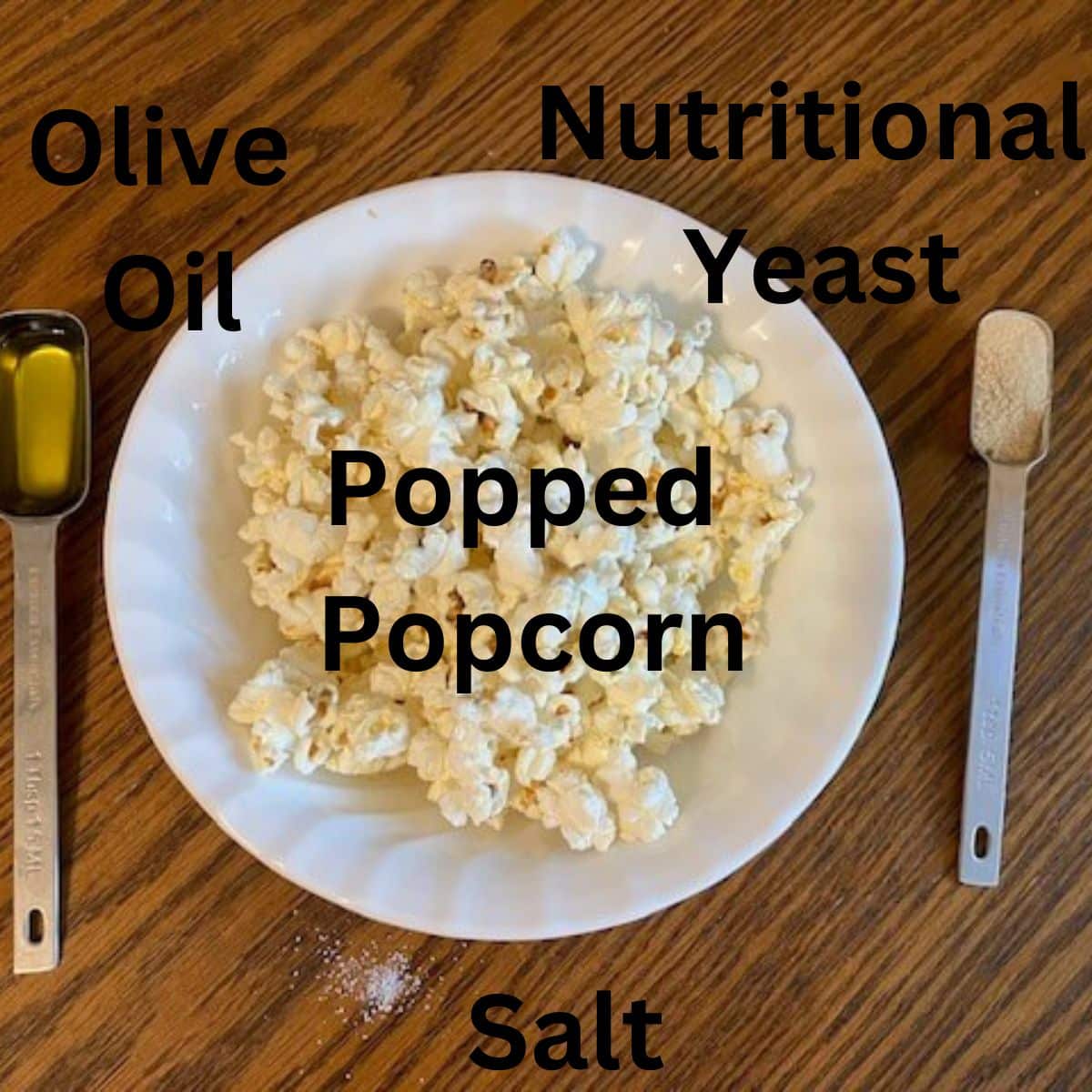 ingredients for the vegan popcorn recipe (popped popcorn, nutritional yeast, olive oil, and salt) with labels on a wood table.
